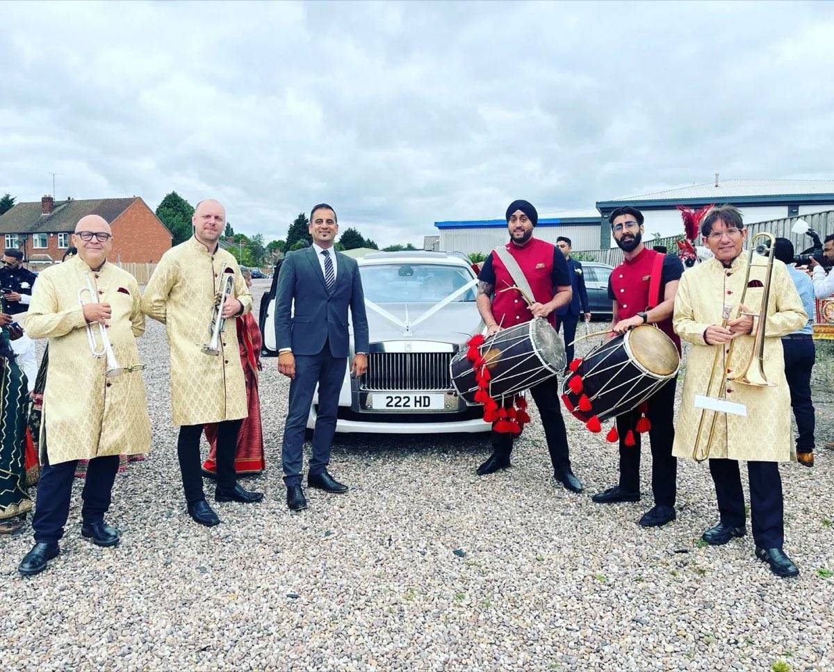 Band baja musicians with trumpets, trombone and dhol drums beside a white Rolls-Royce at a wedding baraat